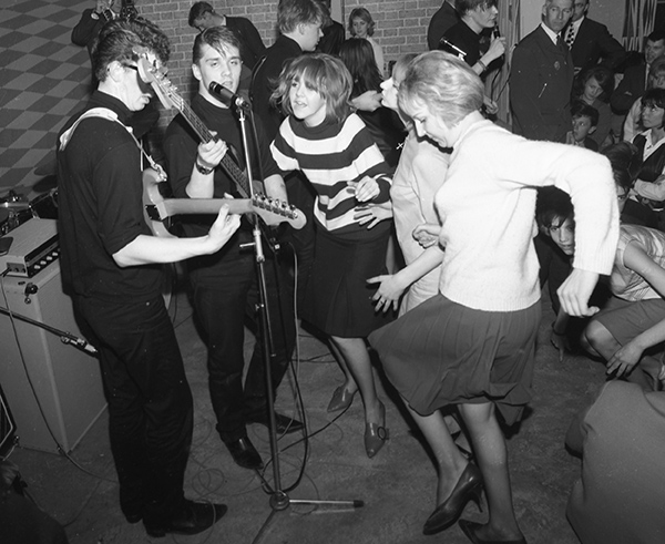 Två killar i ett band spelar gitarr och tjejer som dansar bredvid.