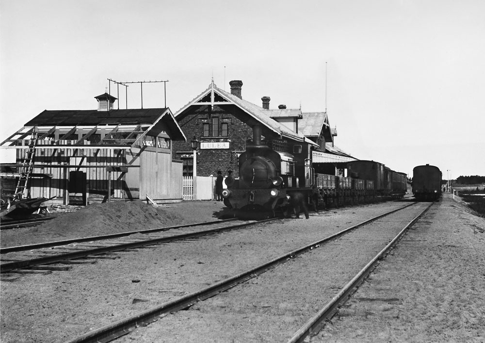 Ånglok med vagnar stående vid järnvägsstation.