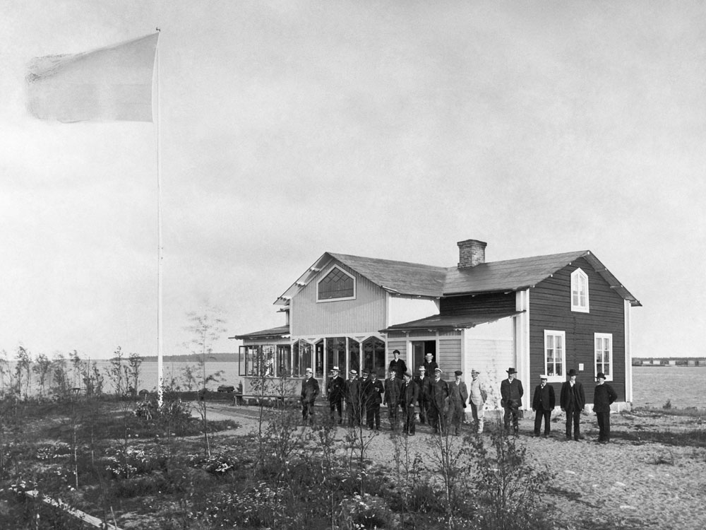 Trähus på skärgårdsö med män på lång rad framför. Flaggstång med flagga intill.