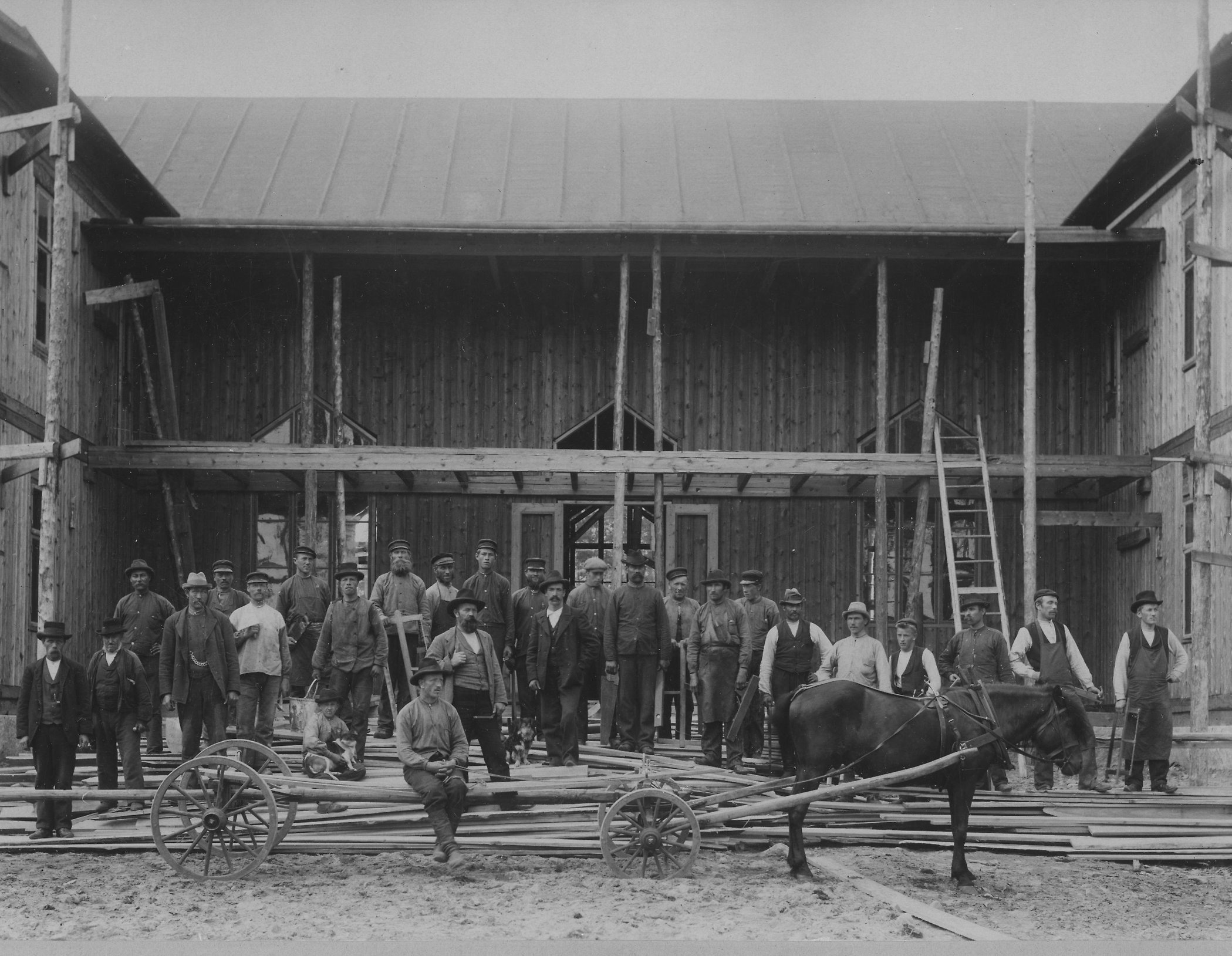 Svartvit bild från 1900 som visar flera personer framför en stor träbyggnad. Byggnaden håller på att uppföras.